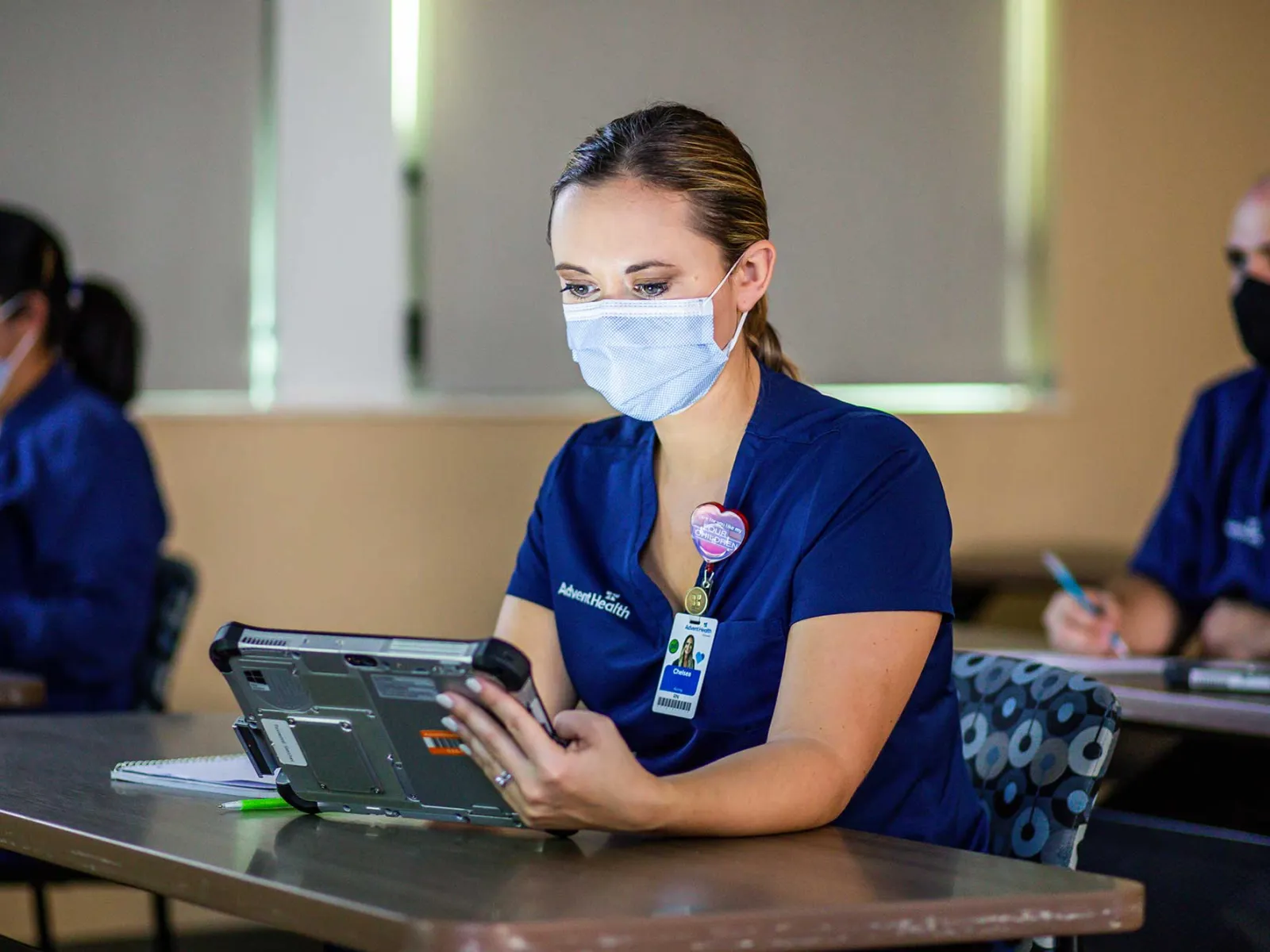 Woman in a surgical mask sitting at a desk with a tablet.