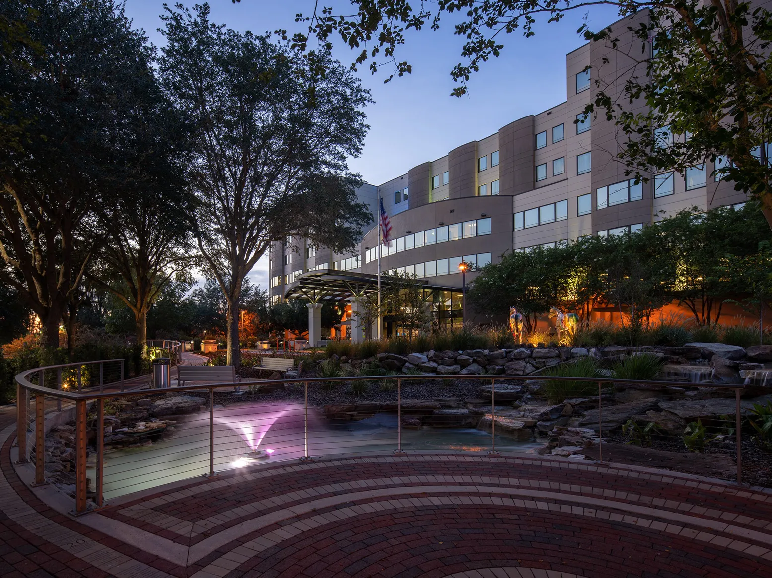 A photo of AdventHealth Ocala taken at dusk. A brick walking path with a railing is in the foreground and the hospital is in the background.