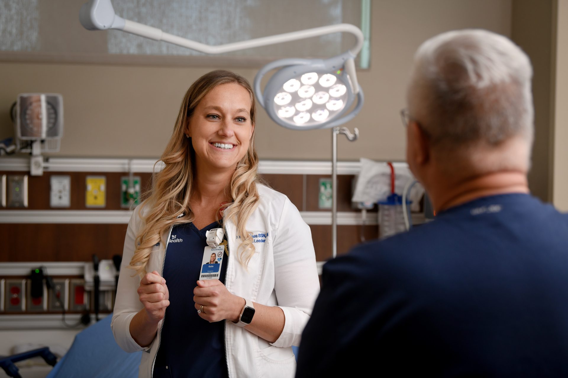A healthcare professional in a white coat is speaking with a patient in a hospital room. Medical equipment and a bed are visible in the background.