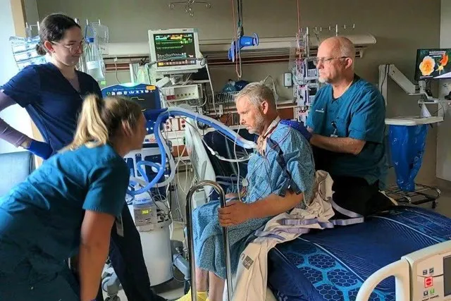 Patient seated in a wheelchair with family member and clinicians participating in a music activity in a hospital room.